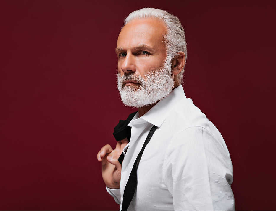 Confident silver-haired man in a white shirt holding a black tie against a burgundy background